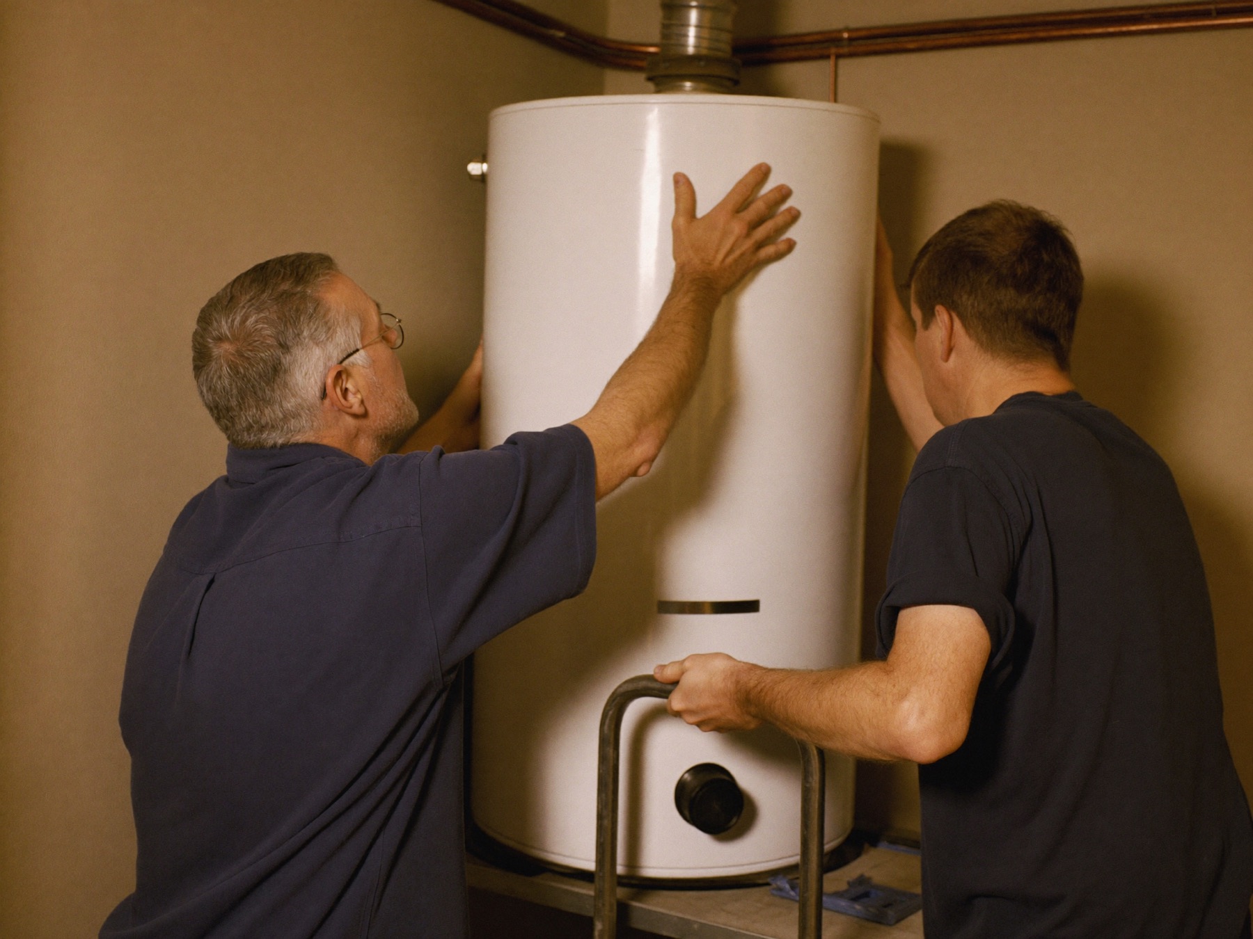 Two Hartwell plumbers moving a new tank water heater into place on a hand truck