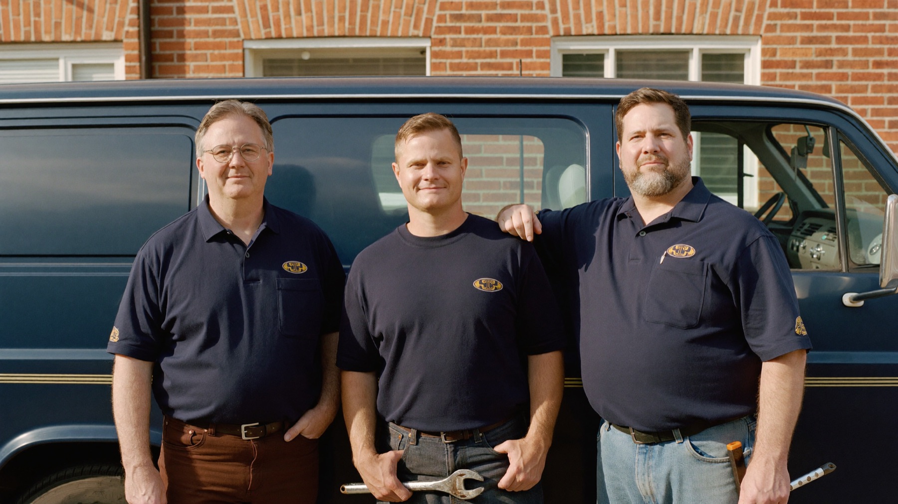 The Hartwell crew — Dan, Pete, and Ray in navy work shirts — standing with the navy van outside a brick west-side house